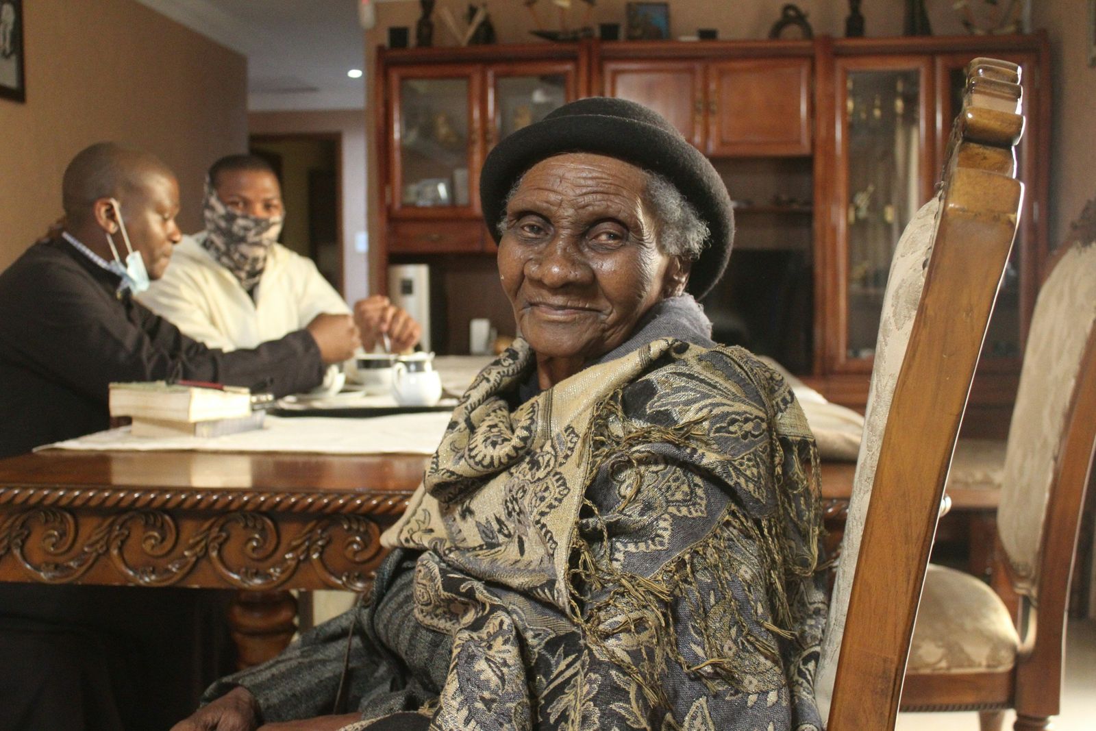 A smiling older Black woman in a patterned jacket and hat seated at a table in a warm community room