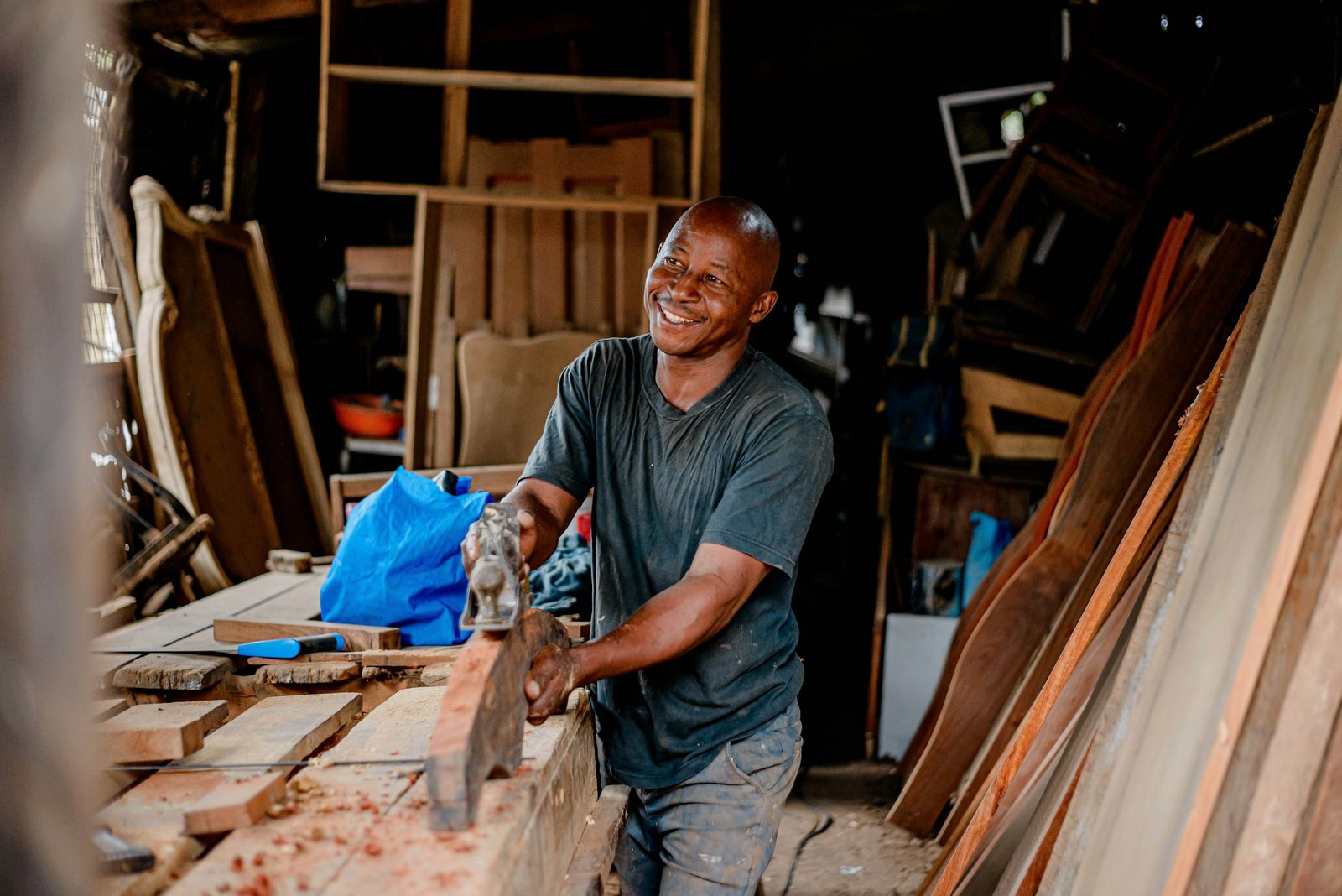 A smiling Black tradesman planing a wooden board in a workshop filled with furniture and lumber